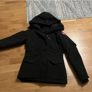 Svart dunjacka från Canada Goose XS - Säljer en svart dunjacka från Canada Goose i storlek XS. Jackan har huva, dragkedja och fickor med lock framtill samt klassisk Canada Goose-logga på ärmen. Perfekt för kalla dagar med sin varma och stilrena design. Kan skcika via post nord med. 