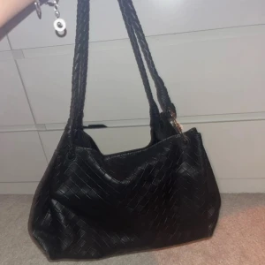 Svart flätad axelväska med innerficka - Snygg svart axelväska i flätad skinnimitation med två rejäla handtag. Väskan har magnetlås och en matchande innerficka med dragkedja. Perfekt storlek för att få plats med allt du behöver. Stilren och trendig design som lyfter varje outfit.