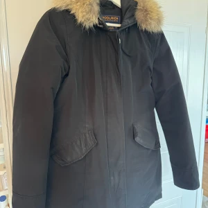 Svart parkajacka från Woolrich med päls - Säljer en svart parkajacka från Woolrich med huva och snygg pälsdetalj runt kragen. Jackan har två stora fickor framtill och stängs med dragkedja och knappar. Perfekt för kalla dagar och riktigt stilren look.