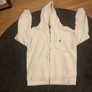 Vit hoodie från Polo Ralph Lauren - Snygg vit hoodie från Polo Ralph Lauren med dragkedja och klassisk logga broderad på bröstet. Tröjan har huva med snörning, känguruficka och är tillverkad i mjuk bomull. Perfekt för en chill och stilren look.