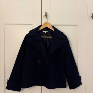 Mörkblå skepparkavaj från Design by Si - Stilren mörkblå skepparkavaj från Design by Si i kortare modell. Jackan har dubbelknäppning framtill, bred krage och snygga knapprankor vid ärmsluten. Perfekt för dig som gillar klassisk och tidlös stil. Obs: en knapp saknas på höger armen 