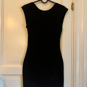 Svart tight klänning med öppen rygg - Snygg svart tight klänning med ärmlös design och öppen rygg. Klänningen har en kort längd och en stilren, minimalistisk look. Perfekt för dig som gillar en klassisk och elegant stil med en twist. Materialet känns mjukt och följsamt mot kroppen.