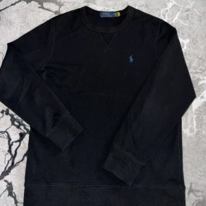 Svart långärmad tröja Polo Ralph Lauren - Svart långärmad tröja från Polo Ralph Lauren med rund halsringning och klassisk blå broderad logga på bröstet. Tröjan är i mjuk bomull och har breda muddar vid ärmslut och nederkant. Perfekt för en stilren och enkel look.