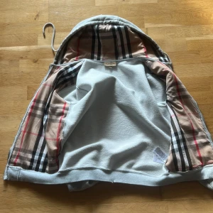 Grå zip-hoodie från Burberry - Snygg grå zip-hoodie från Burberry med klassiskt rutigt foder i huvan och insidan. Jackan har dragsko i huvan, två fickor fram och dragkedja. Perfekt för dig som vill ha en stilren hoodie med lyxig känsla och ikoniska Burberry-detaljer.