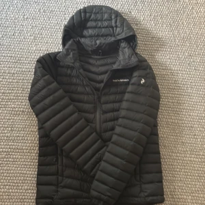 MÖRKGRÖN dunjacka Peak Performance 170 - Mörkgrön dunjacka från Peak Performance med huva och broderad logga på bröstet. Jackan är quiltad med horisontella och snedställda sömmar, har dragkedja framtill och långa ärmar. Perfekt för kyliga dagar och riktigt snygg passform. På ena armen finns det ett pyttelitet hål. 