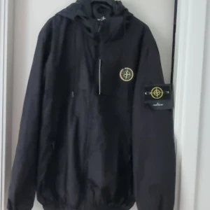Svart vindjacka från Stone Island - Svart vindjacka från Stone Island med ikonisk kompasspatch på ärmen och bröstet. Jackan har huva, dragkedja framtill och elastiska muddar. Oanvänd med alla lappar. 