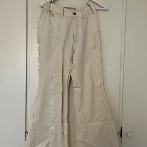 Benvita bootcut jeans med fransdetaljer - Unika benvita jeans med bootcut och råa fransdetaljer längs sidorna. Byxorna har mellanhög midja, bälteshällor och knappstängning. Passar dig med strl 25 i midjan som är under 160cm lång! Har en fläck som ej går bort.