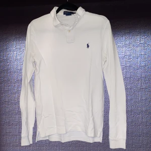 Vit långärmad pikétröja Polo Ralph Lauren - Vit långärmad pikétröja från Polo Ralph Lauren med klassisk krage och två knappar framtill. Tröjan har broderad svart logga på bröstet och är gjord i mjuk bomull. Perfekt för en clean och stilren look.