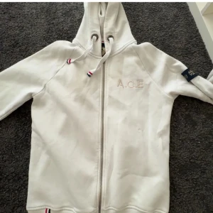 Vit hoodie från WHO'S A.C.E XS - Snygg vit hoodie från WHO'S A.C.E med dragkedja och huva. Tröjan har broderad logga på bröstet, coolt märke på ärmen och detaljer i rött, vitt och blått på snören och insidan av huvan. Tröjan passar även  i storlek S är 177 cm lång och den sitter perfekt,  