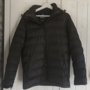 Svart pufferjacka från Tuxer XS - Svart pufferjacka från Tuxer i storlek XS. Jackan har huva, två fickor med dragkedja och quiltad design. Perfekt för kyliga dagar och har en stilren look med diskreta detaljer. Materialet är syntet och jackan stängs med dragkedja framtill.