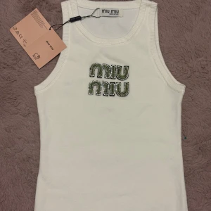Vit linnetopp från Miu Miu - Supersnygg vit linnetopp från Miu Miu med broderad logga framtill i grönt och silverdetaljer. Toppen är ärmlös med ribbade kanter och har en klassisk, rak passform. Perfekt för att lyfta vilken outfit som helst.
