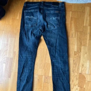 Mörkblå jeans från Nudie Jeans W32 L34 - Säljer ett par mörkblå jeans från Nudie Jeans med klassisk femficksdesign och snygga kontrastsömmar. I storlek W32 L34 Jeansen har raka ben och subtila slitningar för en cool look. Perfekta för dig som gillar stilrena och tidlösa jeans. Model Grim Tim 