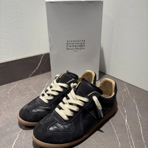 Maison Margiela svarta sneakers - Maison Margiela sneakers i svart skinn och mocka med beige insida och vita snören. Klassisk rund tå och platt gummisula i brunt. Snygg retrostil med kontrasterande material och neutrala färger, perfekt för dig som gillar stilrena och exklusiva sneakers.