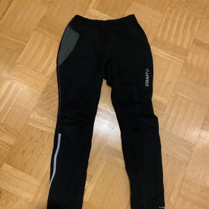 Svarta träningsbyxor från Craft - Svarta träningsbyxor från Craft med grå detalj på ena sidan och reflekterande inslag längs benet. Byxorna har smal passform och är tillverkade i syntetmaterial, perfekta för löpning eller annan träning. Craft-logga på låret.