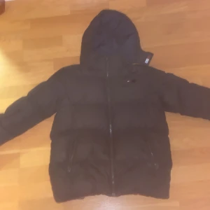 Svart pufferjacka Tommy Hilfiger 152 - Svart pufferjacka från Tommy Hilfiger med huva och dragkedja framtill. Jackan har två fickor med dragkedja och logga på bröstet samt insidan av kragen. Perfekt för kalla dagar och riktigt snygg streetstil.