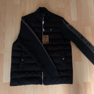 Svart dunväst från Burberry, storlek L - Snygg svart dunväst från Burberry med klassiskt rutmönster på ryggen och logotyp på bröstet. Västen har quiltad framsida, två fickor med knapp och dragkedja framtill. Materialet är polyester och dun, med ribbade detaljer och Burberry-tryck på axlarna.