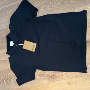 Svart pikétröja från Burberry - Svart pikétröja från Burberry med klassisk krage och knappslå. Tröjan har diskret broderad logga på bröstet och är gjord i bomull. Perfekt för en clean och stilren look.