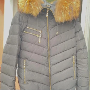 Svart pufferjacka med päls från Hollies - Säljer en svart pufferjacka från Hollies med stor fluffig päls på huvan. Jackan har guldfärgade dragkedjor, flera fickor och snygga quiltade detaljer. Perfekt för dig som vill ha en varm och stilren vinterjacka med lyxig känsla.