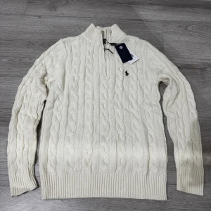 Kabelstickad vit tröja från Polo Ralph Lauren  - Kabelstickad vit tröja från Polo Ralph Lauren med halv dragkedja och hög krage. Klassisk broderad logga på bröstet. Tröjan är i mjuk ullblandning och har ribbade muddar och nederkant. Perfekt för lager-på-lager under kyliga dagar.