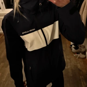 Marinblå och vit seglarjacka från Henri Lloyd - Snygg vindjacka från Henri Lloyd i svart med vita detaljer och logga på bröstet och ryggen. Jackan har huva med dragsko, hel dragkedja och praktiska fickor. Perfekt för blåsiga dagar och stilren look.