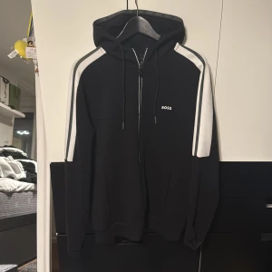Svart hoodie från Hugo boss - Svart hoodie från BOSS med vita ränder längs axlarna och ärmarna. Tröjan har huva med snörning, dragkedja framtill och diskret BOSS-logga på bröstet. Perfekt för en sportig och stilren look. Aldrig använd nyskick!