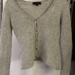 Grå fluffig kofta från Twilfit XS - Supermjuk och fluffig grå kofta från Twilfit i storlek XS. Koftan har v-ringning, långa ärmar och knappar framtill. Materialet är stickat och ger en mysig känsla, perfekt för kyliga dagar. Enkel och stilren design som är lätt att matcha med dina favoritjeans.