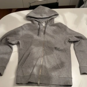 Grå hoodie från Replay med dragkedja - Säljer en grå hoodie från Replay med mjukt fleecefoder och dragkedja framtill. Tröjan har huva med snörning, två fickor och Replay-logga på bröstet.  Nytt skick 
