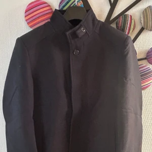 Rock - Stilren svart rock med hög ståkrage och knappstängning framtill. Jackan har en rak och klassisk siluett som ger en clean look. Perfekt för dig som gillar minimalistisk stil och vill ha något som funkar till många outfits. Materialet känns tjockt och varmt. Aldrig använd då jag råkade beställa fel storlek