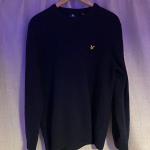 Mörkblå stickad tröja från Lyle & Scott - Snygg mörkblå stickad tröja från Lyle & Scott med klassisk rund hals och gul broderad logga på bröstet. Tröjan har långärmad passform och ribbade muddar vid ärmslut och nederkant. Perfekt för dig som gillar stilrena och enkla plagg.