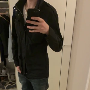 Svart field jacka från J.Lindeberg - Svart fieldjacka från J.Lindeberg med hög krage, fyra stora fickor med lock och knappar, samt snygga metallknappar och logodetalj på bröstet. Jackan har dragkedja och är tillverkad i slitstark polyester, perfekt för kyliga dagar.