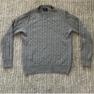 Grå kabelstickad tröja från Gant - Kabelstickad grå tröja från Gant i premium bomull. Klassisk rund halsringning, ribbade muddar och broderad Gant-logga med sköld på bröstet. Perfekt för lager-på-lager och snygg till jeans eller chinos.