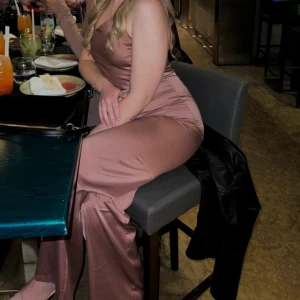 Rosa jumpsuit med smala axelband - Säljer en elegant jumpsuit med smala axelband och figurnära passform. Klänningen har en slät yta och lätt glansigt material som ger en lyxig känsla. 