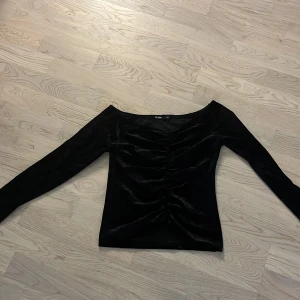 Svart sammets topp från BikBok - Säljer en svart sammets topp från BikBok i storlek XS. Toppen har lång ärm, v-ringad hals och snygg rynkdetalj framtill. Materialet är mjukt och glansigt, vilket ger en lyxig känsla. Perfekt för dig som gillar trendiga och stilrena plagg.