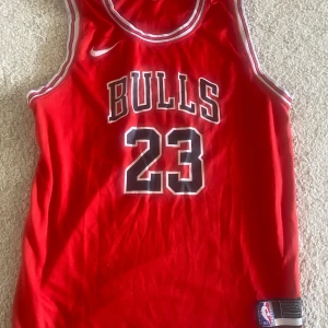 Chicago Bulls Jordan 23 baskettröja - Säljer en röd Chicago Bulls baskettröja från Nike med nummer 23 och Jordan på ryggen. Tröjan är ärmlös och har vita och svarta detaljer vid krage och ärmhål. Materialet är lätt och ventilerande polyester, perfekt för sport eller streetwear.