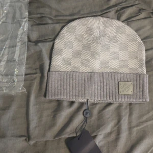 Grå Louis Vuitton rutig mössa - Snygg grå mössa från Louis Vuitton med klassiskt rutigt mönster och ribbad uppvikt kant. Mössan har en diskret LV-logga på läderpatch framtill och är tillverkad i mjukt material som känns lyxigt. Perfekt accessoar för att lyfta din vinterstil.