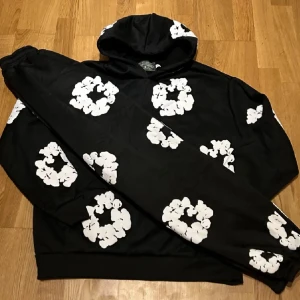 Denim tears hoodie med byxor  - Svart hoodie med huva och vita blommönster över hela tröjan. Tröjan har en klassisk passform, långa ärmar och känguruficka framtill. Perfekt för dig som gillar streetwear och vill sticka ut med en cool design.