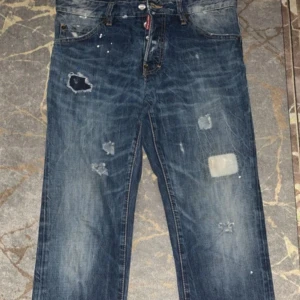 Dsquared2 blå jeans med patchar - Snygga blå jeans från Dsquared2 med slitningar, lagade hål och patchar på benen. Klassisk femficksmodell med läderpatch bak i midjan. Jeansen har en rak passform och coolt distressed utseende. Tillverkade i Italien av bomull.