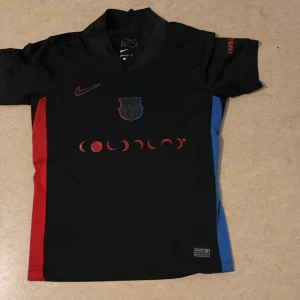 Svart Nike FC Barcelona fotbollströja - Unik svart FC Barcelona fotbollströja från Nike med korta ärmar. Tröjan har röda och blå detaljer längs sidorna, klubbmärke på bröstet och Nike-logga i rödblå gradient. Tillverkad i Dri-FIT material som andas, perfekt för match eller träning.