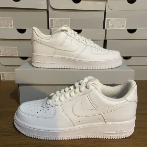 Nike Air Force 1 helvita sneakers - Klassiska Nike Air Force 1 sneakers i helvitt skinn. Modellen har låg profil, rund tå, perforeringar på tån för extra ventilation och tydlig Nike swoosh på sidorna. Tjock vit sula och snörning framtill. Perfekta för en clean och stilren look.