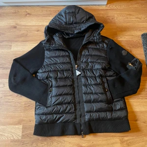 Svart Moncler pufferjacka med huva - Svart pufferjacka från Moncler med stickade ärmar och huva. Jackan har dragkedja framtill, en ficka med dragkedja på ärmen och klassisk Moncler-logga. Materialmix av polyester och stickat ger en modern vibe. Perfekt för kyliga dagar när du vill hålla stilen. Jackan är inte äkta men den har NFC taggen på logan vid armen. Jackan har ett litet hål vid ärmen.