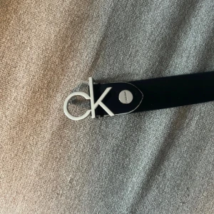 Svart skärp från Calvin Klein i skinn - Snyggt svart bälte från Calvin Klein i äkta skinn med stor silvrig CK-logga som spänne. Bältet har Calvin Klein-tryck längs remmen och är perfekt för att lyfta vilken outfit som helst. Klassisk och stilren design som passar till jeans eller kostymbyxor.