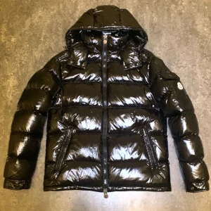 Svart Moncler Maya dunjacka med huva - Säljer en svart, glansig Moncler Maya dunjacka med huva och dragkedja. Jackan har två fickor med dragkedja framtill, klassisk Moncler-logga på ärmen och quiltad design. Perfekt för kalla dagar och riktigt snygg streetstil.