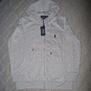 Grå hoodie från Polo Ralph Lauren - (Kan passa M) Snygg ljusgrå hoodie från Polo Ralph Lauren med dragkedja och huva. Klassisk logga broderad på bröstet och praktiska fickor framtill. Perfekt för en chill och stilren look.