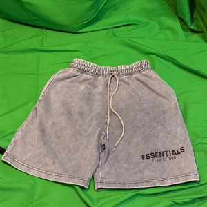Grå shorts Essentials Fear of God - Säljer ett par grå shorts från Essentials Fear of God med tvättad look. De har elastisk midja med snörning, sidofickor och en bakficka. Loggan är tryckt framtill. Perfekta för chill dagar och streetwear-stil.Liknande modell som originalet!