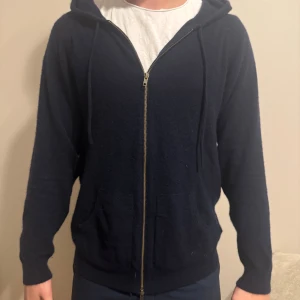 Mörkblå hoodie i kashmir från Davida - Snygg mörkblå hoodie från Davida i 100% kashmir. Tröjan har dragkedja framtill, huva med snörning och två fickor. Långärmad och supermjuk, perfekt för chill dagar. Stilren design som funkar till jeans eller mjukisbyxor.