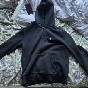 Svart hoodie från Polo Ralph Lauren - Svart hoodie från Polo Ralph Lauren. Tröjan har huva med snörning, dragkedja framtill och en stor magficka. Tillverkad i mjuk bomull och har långärmad passform. Perfekt för chill dagar och enkel att matcha med jeans.