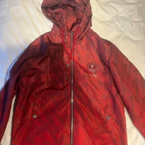 Trapstar windbreaker - Säljer en röd vindjacka från Trapstar med camomönster, broderad logga på ryggen och patch på bröstet. Jackan har huva med snörning, dragkedja framtill och två fickor med tryckknappar. Perfekt för dig som vill sticka ut med streetwear-vibe.