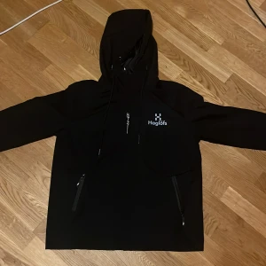 Svart vindjacka från Haglöfs med huva - Svart vindjacka från Haglöfs med stor logga på ryggen och mindre logga på bröstet. Jackan har huva med snörning, två snedställda fickor med dragkedja och en clean, sportig look. Perfekt för dig som gillar outdoor och streetstyle Storlek L.