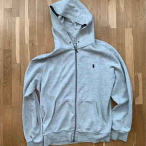 Ljusgrå hoodie från Polo Ralph Lauren - Snygg ljusgrå hoodie från Polo Ralph Lauren med dragkedja och klassisk logga på bröstet. Tröjan har huva, två fickor framtill och är tillverkad i mjuk bomullsmix. Perfekt för en chill och stilren look.