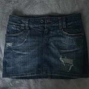 Mörkblå jeanskjol med slitningar och hjärtdetalj - Säljer en mörkblå jeanskjol från only med coola slitningar framtill och snygga bakfickor. Kjolen har klassisk knapp och dragkedja, samt en hjärtdetalj på bakfickan. Lågmidjad och lagom kort